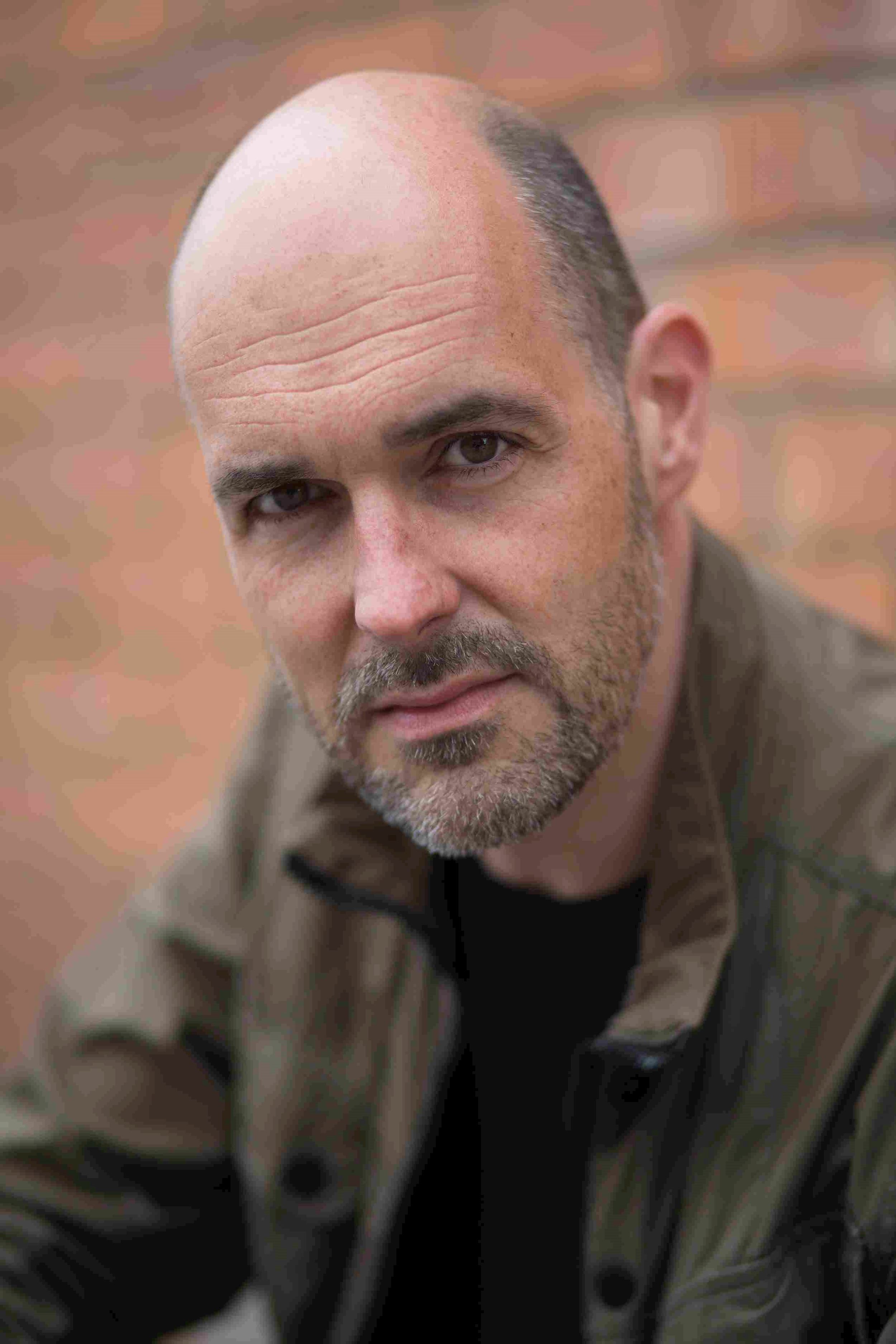 James McLean: Man with a close-cropped beard and shaved head, wearing a dark green jacket, gazing seriously into the camera with a brick wall in the background.