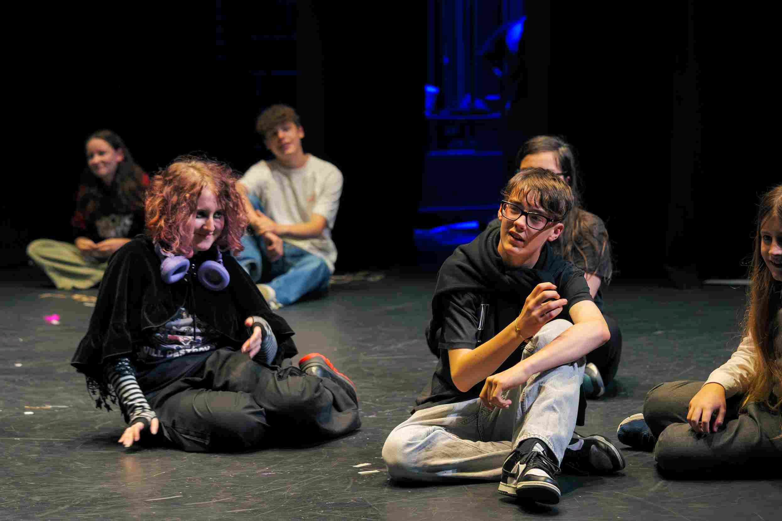 Young Company participants sit on the stage floor in Studio 2 during a rehearsal or workshop. A group of teenagers are engaged in conversation, with one person gesturing animatedly while others listen and smile.