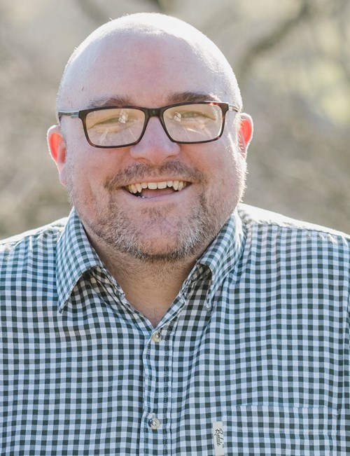 Headshot of Pete Toon. He has a shaved head, glasses, and a beard, wearing a checked shirt, smiling warmly at the camera.