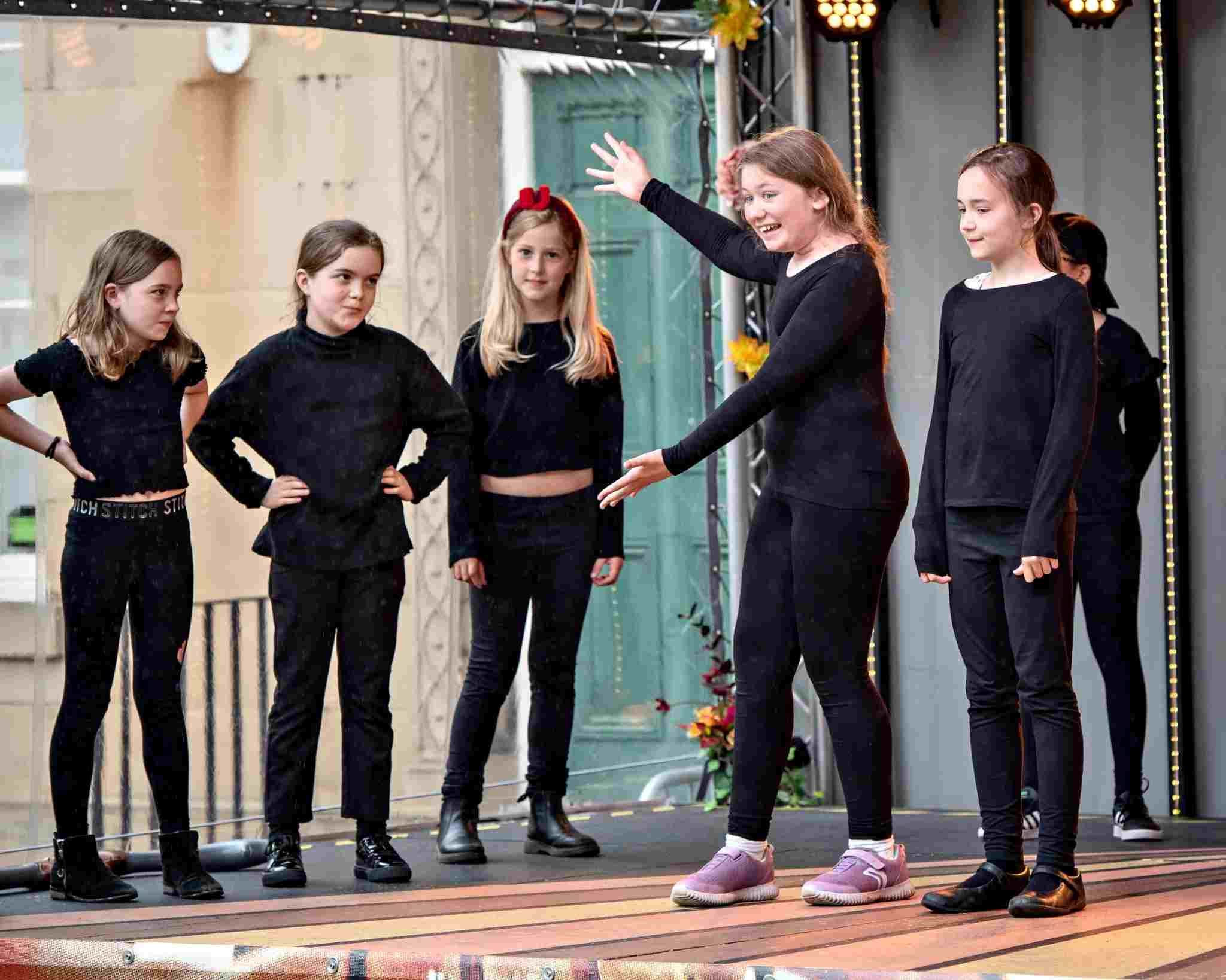 A group of six young girls perform on an outdoor stage, all dressed in black. One girl stands in the centre with arms outstretched, smiling animatedly, while the others watch her with interest. Stage lights and a decorative backdrop are visible.