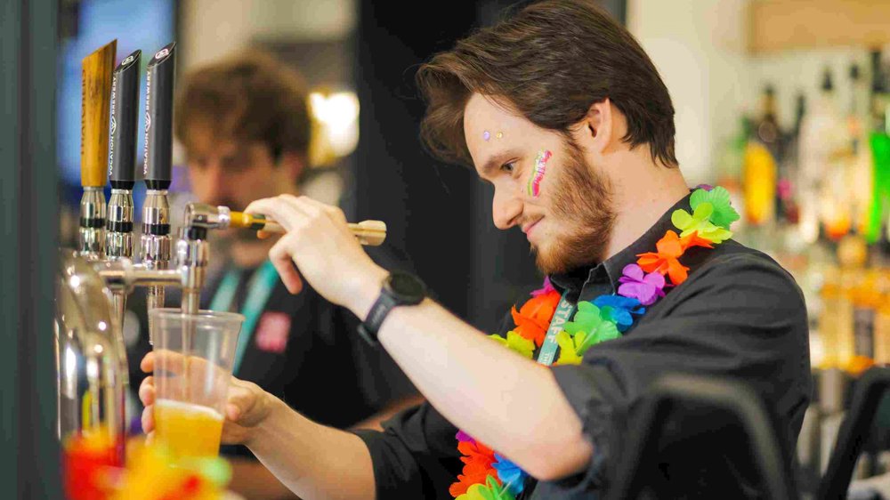A Lawrence Batley Theatre team member pours a drink at the bar, wearing a colourful flower garland and face paint, with a focused expression. The bar is lively and well-stocked, with another staff member visible in the background.