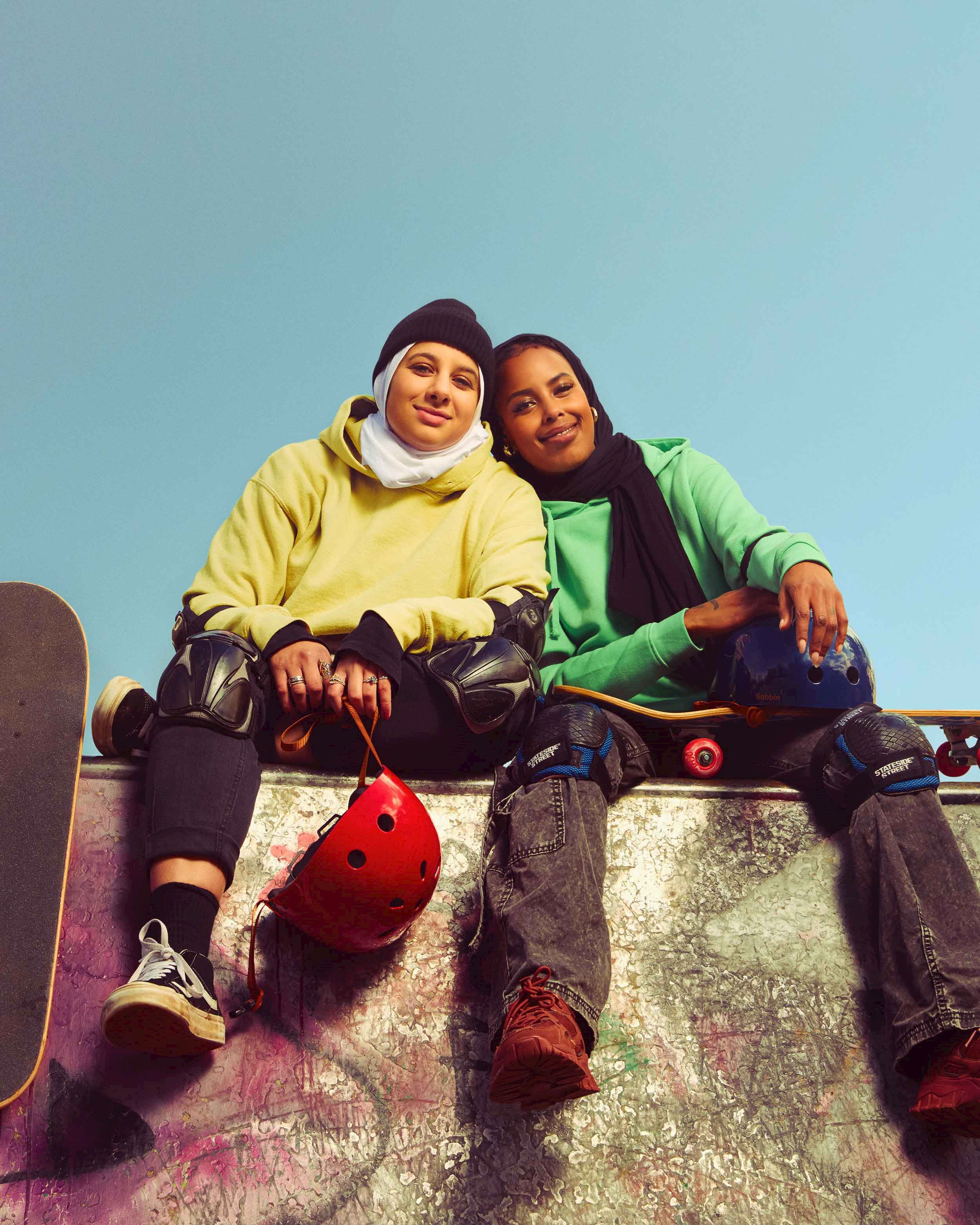 An image of two young women in hoodies and headscarves sitting on a skate ramp with skateboards and helmets, smiling.