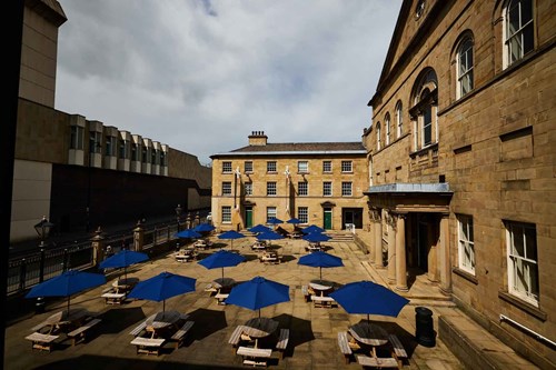 An image of Lawrence Batley Theatre’s outdoor courtyard. The courtyard is filled with wooden picnic tables, each shaded by a large blue umbrella, arranged in an orderly fashion.