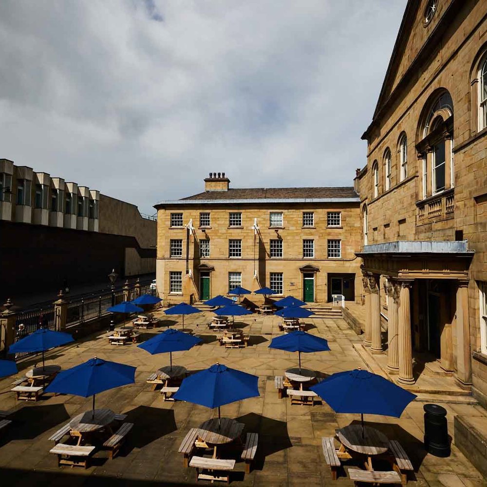 An image of Lawrence Batley Theatre’s outdoor courtyard. The courtyard is filled with wooden picnic tables, each shaded by a large blue umbrella, arranged in an orderly fashion.