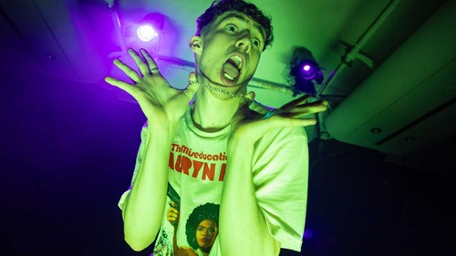 A young man is making a playful and exaggerated expression, holding his necklace with both hands. He is wearing a T-shirt that reads 'The Miseducation of Lauryn Hill' and is bathed in green stage lighting.