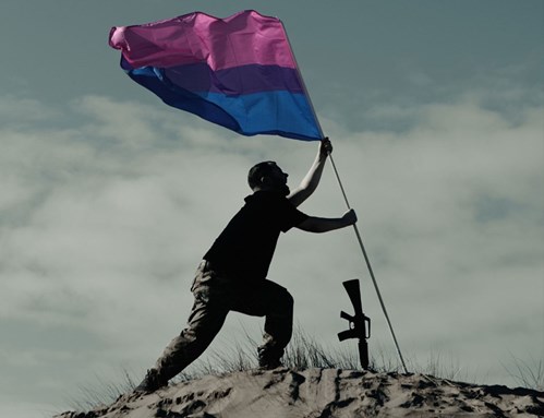 A man wearing army trousers and a black t-shirt stands holding a bisexual pride flag on top of a sand dune. 