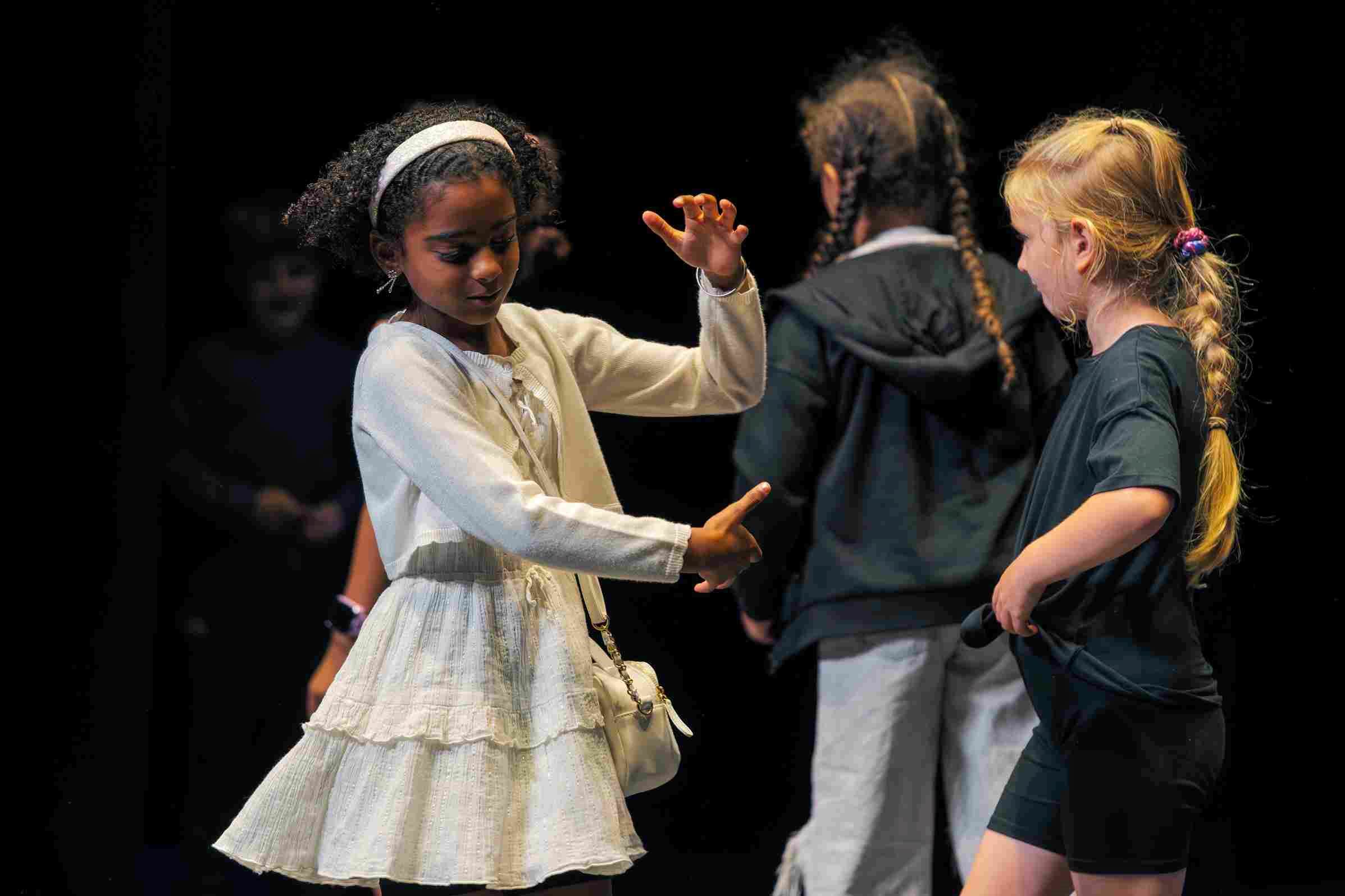 Members of the Stage Door group from the Young Company youth theatre rehearse on stage. A girl in a white dress gestures expressively with her hands while another girl in black shorts and a t-shirt watches closely.