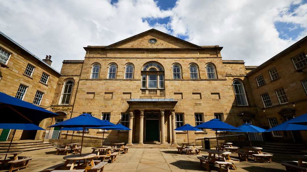 Picture of Lawrence Batley Theatre, which is a Georgian stone building set back behind a courtyard with wooden tables and benches, with blue umbrellas, under a blue cloudy sky in Huddersfield