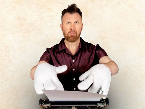 Comedian Jason Byrne wears a silk, red shirt. His eyebrows are raised as he pulls a thinking face. He has over-sized, white cartoon hands on and is trying to type on an old fashion typewriter. 