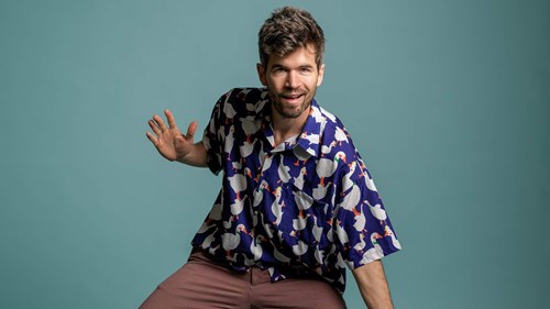 Comedian Ivo Graham with dark hair and a beard wearing a light blue shirt with lots of white ducks patterned on it. 
