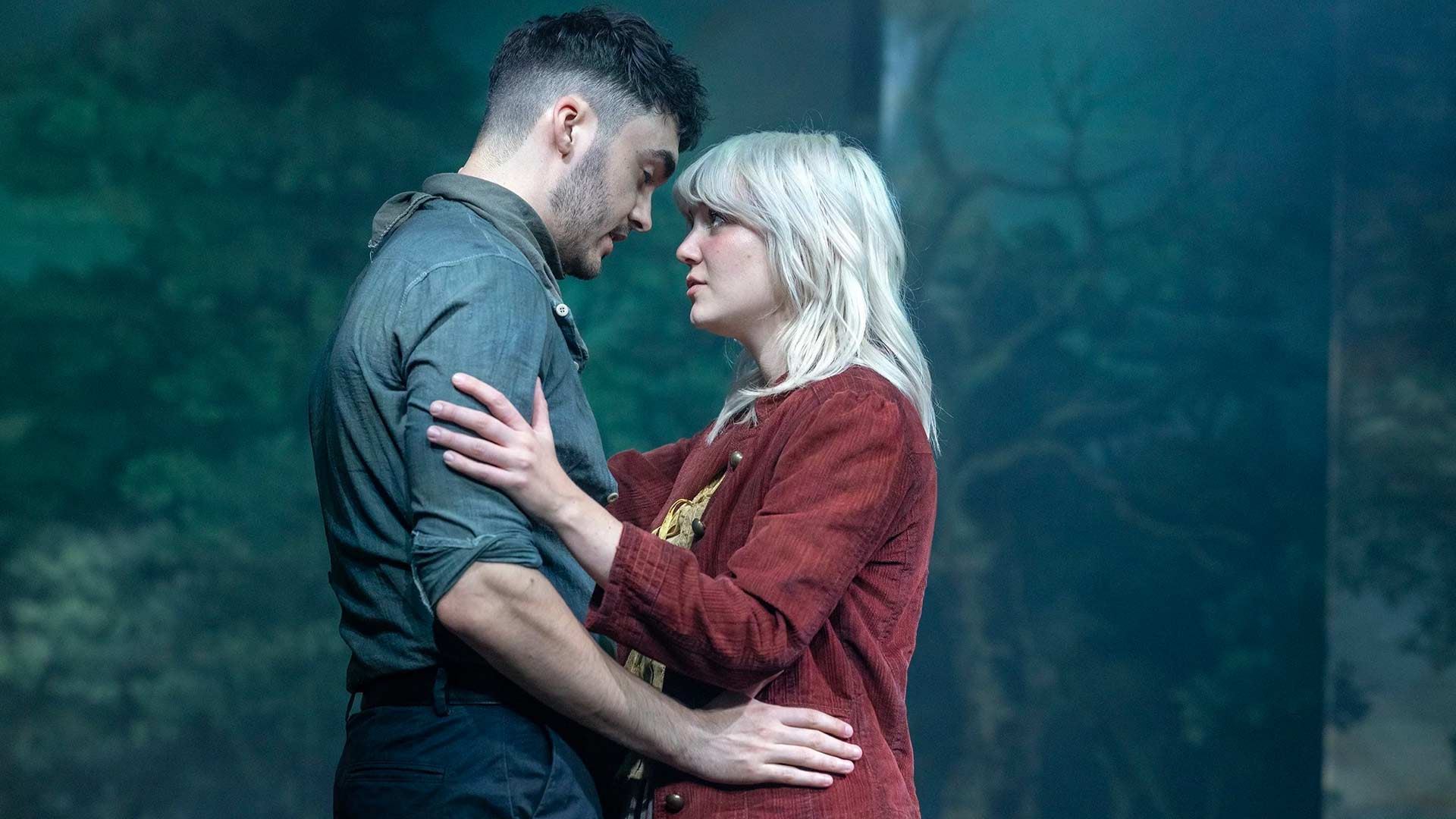 Two people embrace and gaze at each other on a forest-themed stage, creating an intimate, emotional moment.