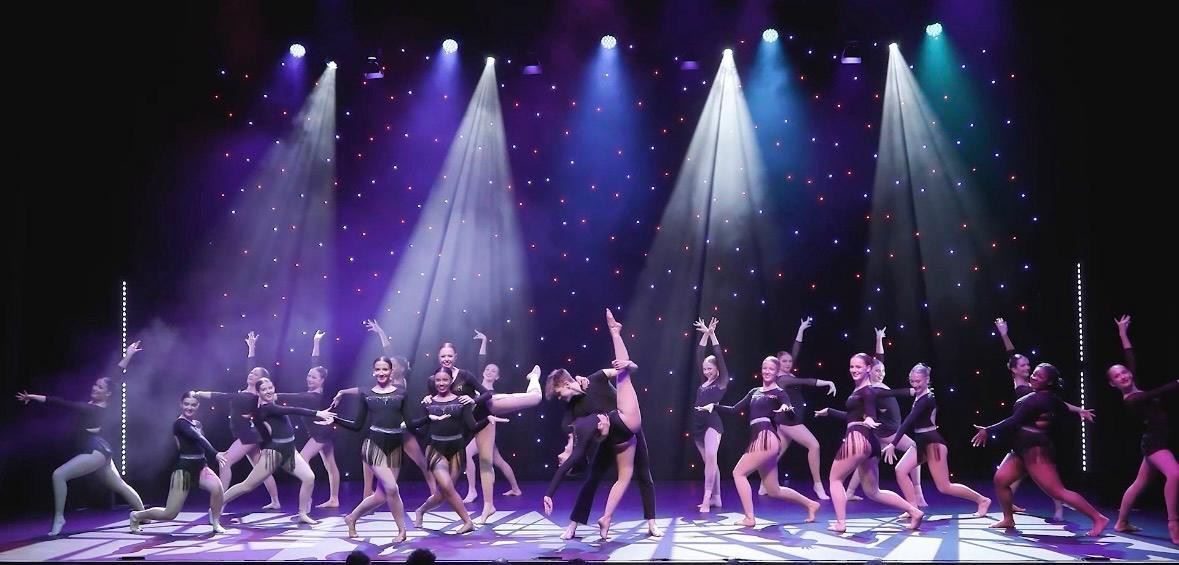 A large group of dancers perform on stage under bright spotlights, wearing coordinated dark costumes. They strike expressive poses and lifts against a starry backdrop, creating a dynamic, high-energy performance scene.
