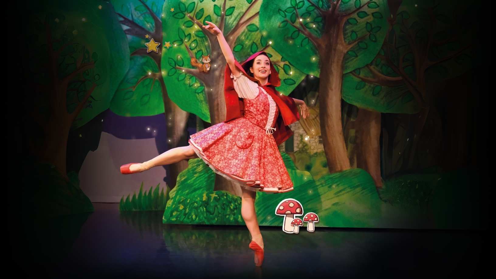 A ballet dancer dressed as Little Red Riding Hood performs a graceful pose on stage, surrounded by a colourful forest backdrop with illustrated trees, stars, and toadstools.