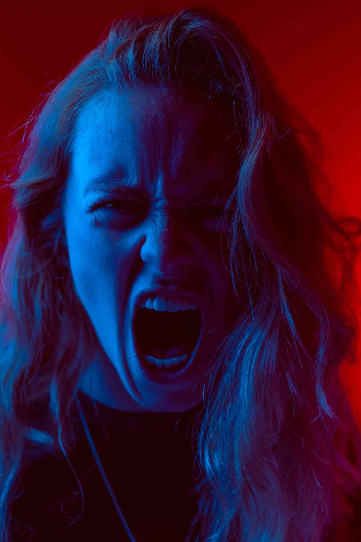 A person with long hair bathed in dramatic red and blue lighting, screaming or shouting with intense emotion, against a vivid red background.