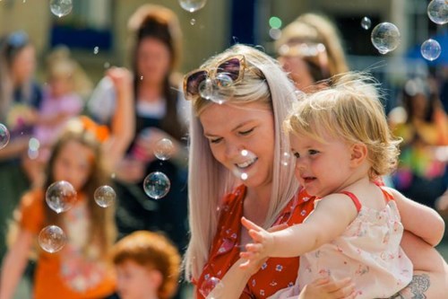 A cheerful outdoor scene showing a woman in a red dress holding a toddler, surrounded by floating bubbles. The woman and child are smiling as the child reaches out to touch the bubbles, with a lively group of people blurred in the background.
