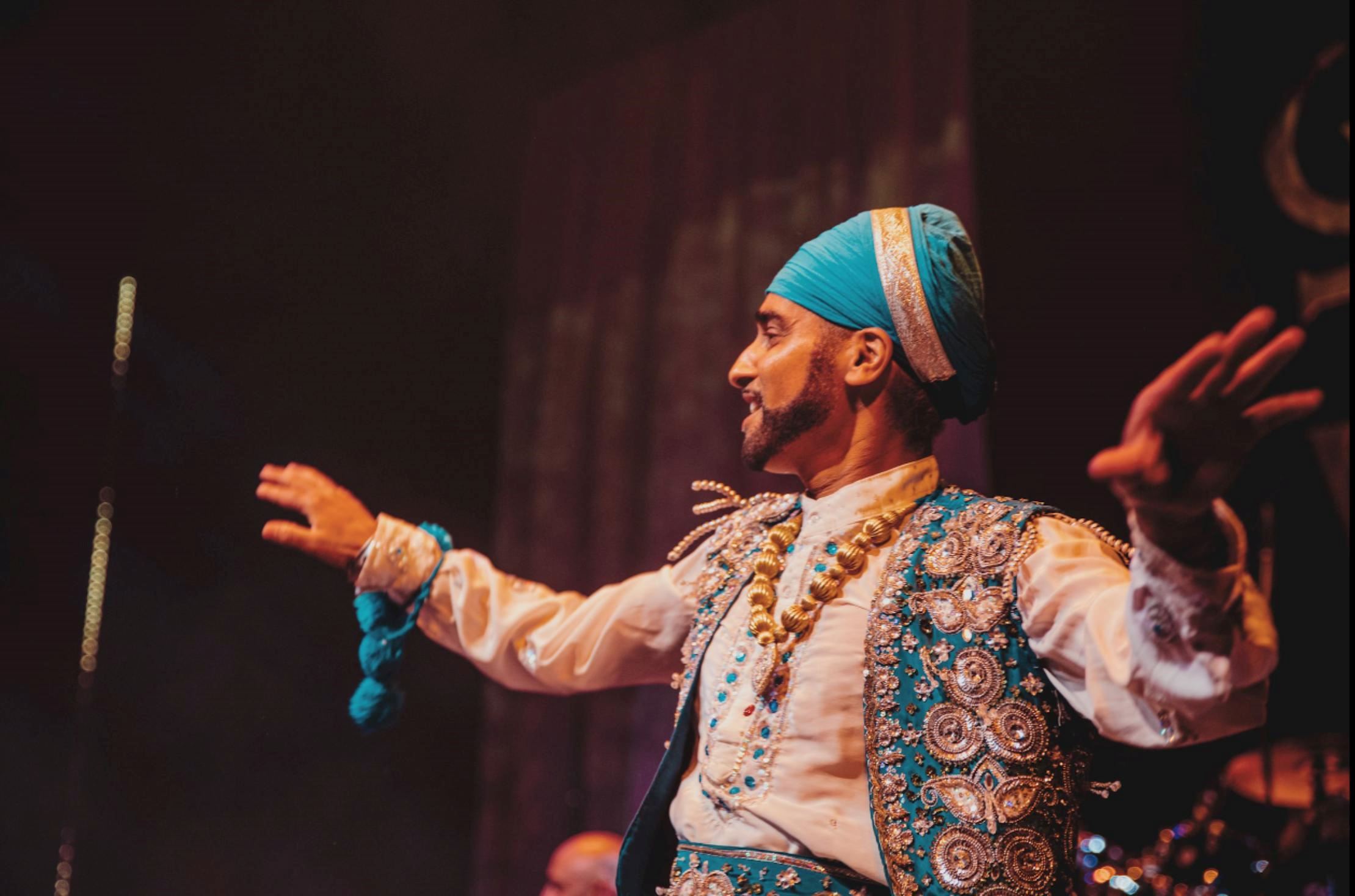 A performer in a blue and gold traditional costume stands on stage with arms outstretched during a live performance, illuminated by warm theatre lighting.