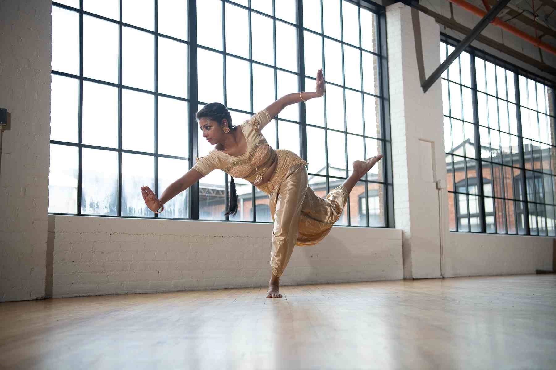A dancer in a gold traditional Indian costume balances gracefully on one foot, arms extended and one leg raised behind her, in a bright industrial-style studio with large grid windows and wooden floors.