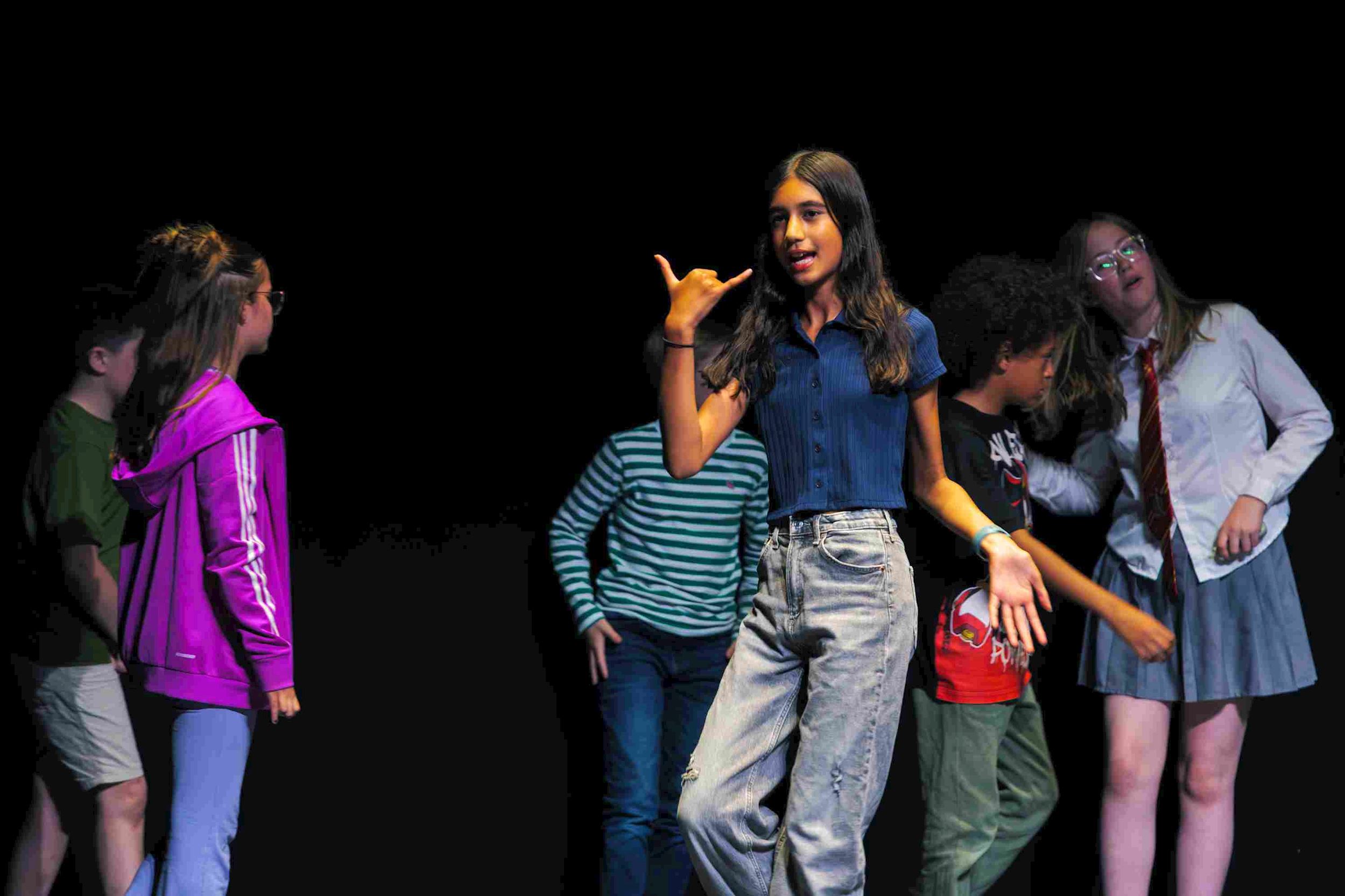 Young performers from the Young Company youth theatre rehearse a scene on stage in Studio 1. A girl in the foreground gestures animatedly with her hands as others stand and interact behind her.