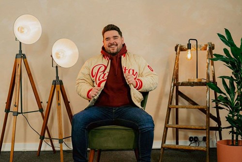 Comedian Adam Rowe smiles at the camera, he is sitting on a green velvet wearing blue jeans, a red jumper and a cream jacket. 