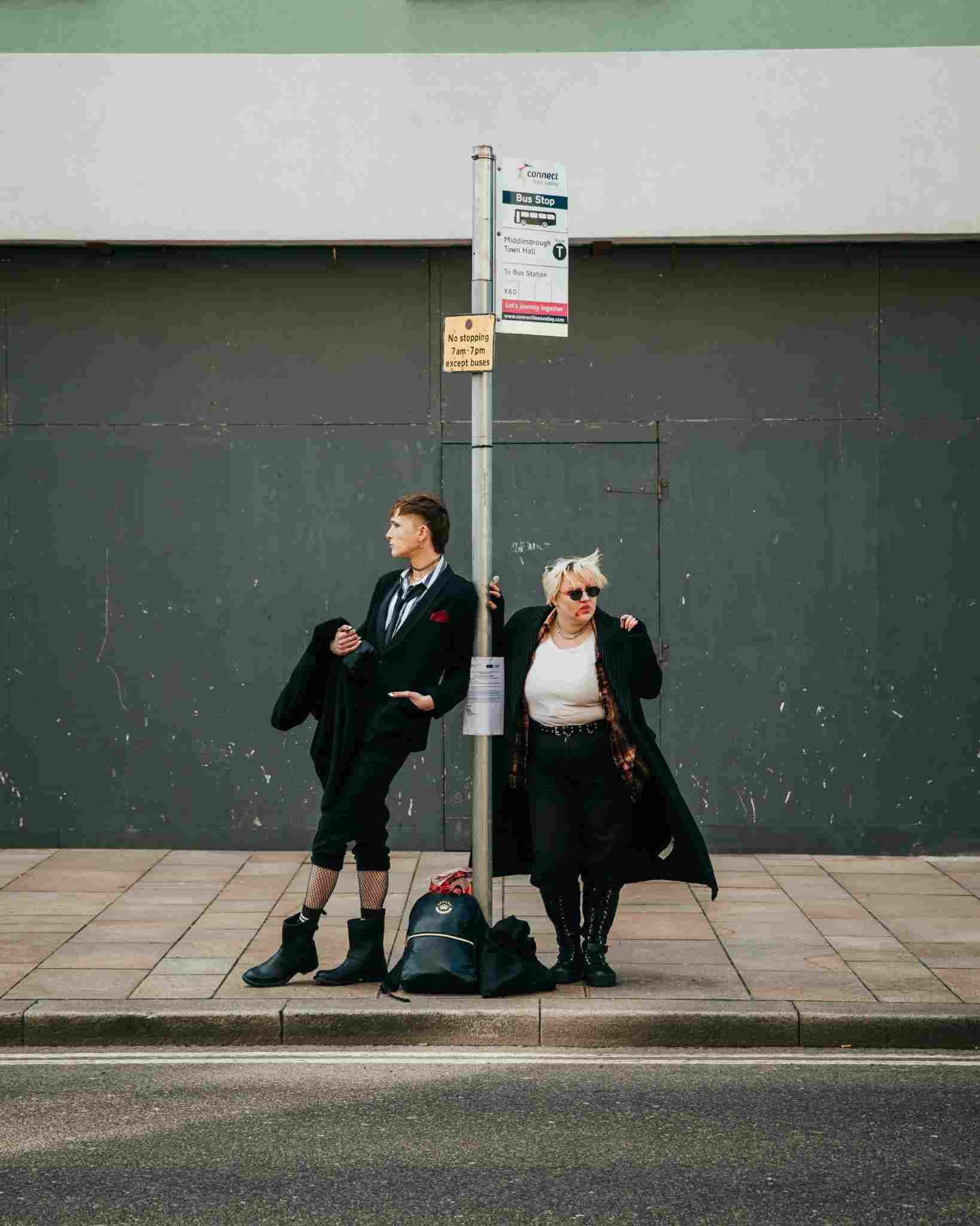 Two people stand at a bus stop on a city pavement, dressed in bold, alternative clothing. One leans against the bus stop pole in a suit and boots, while the other wears sunglasses, a long coat, and checked shirt. A backpack sits on the ground between them against a backdrop of an urban wall.