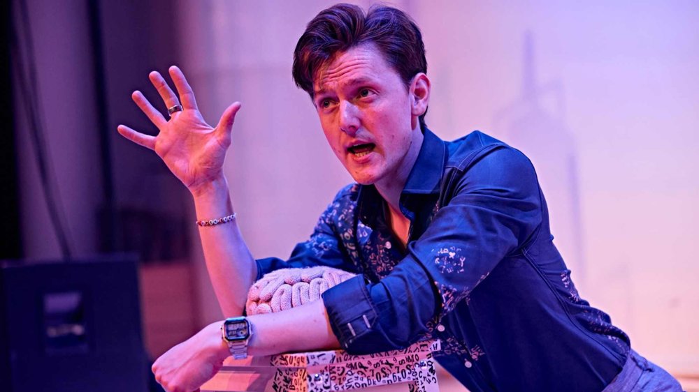 A performer in a blue patterned shirt sits on stage under pink and purple lighting, gesturing expressively with one hand while speaking. Leaning on a stack of props, they appear to be engaged in a storytelling or theatrical performance. The background is softly lit, focusing attention on the performer and their animated expression.