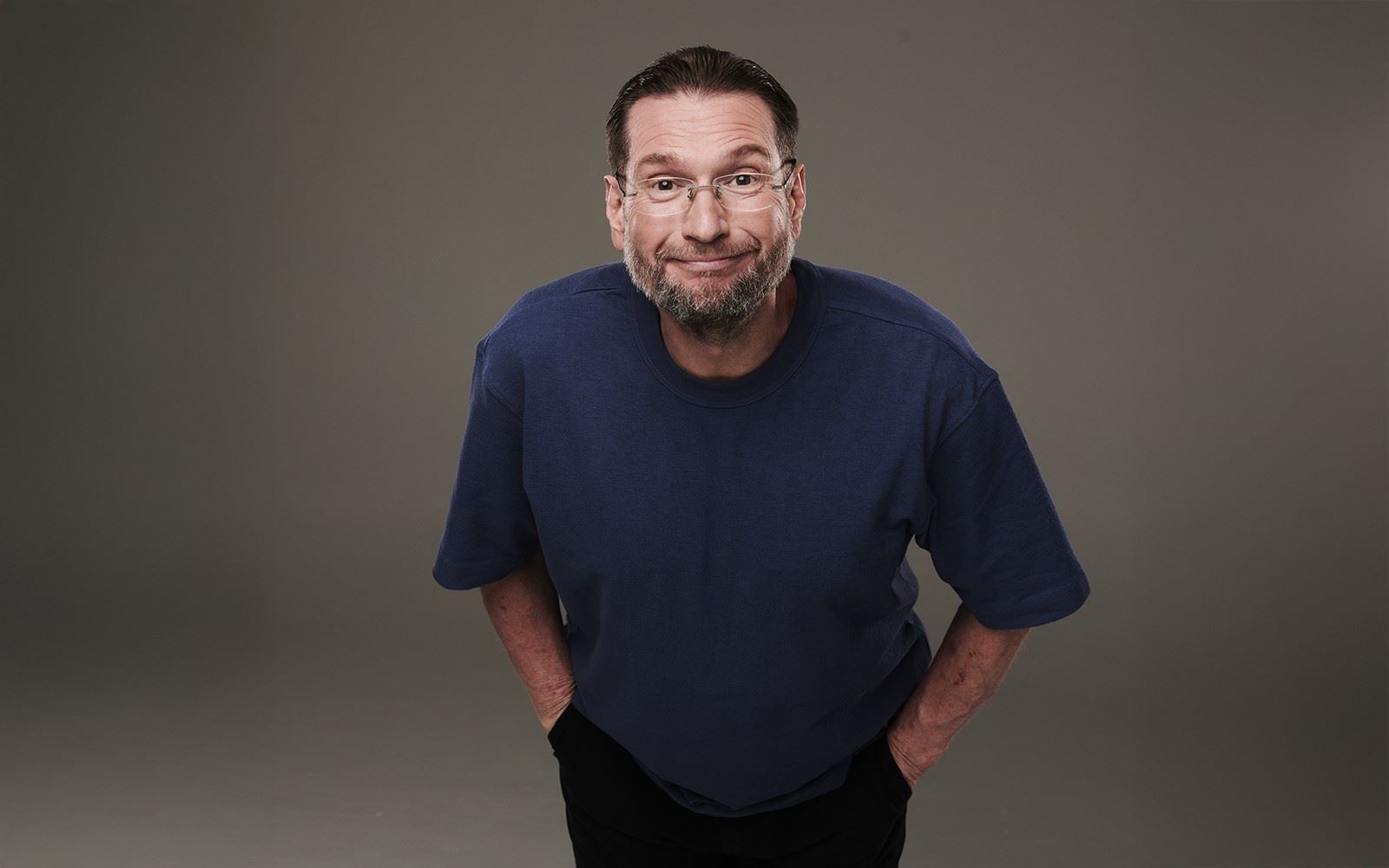 Comedian Gary Delaney stands against a bright blue background, smiling confidently in a dark jacket and shirt, promoting his upcoming tour.