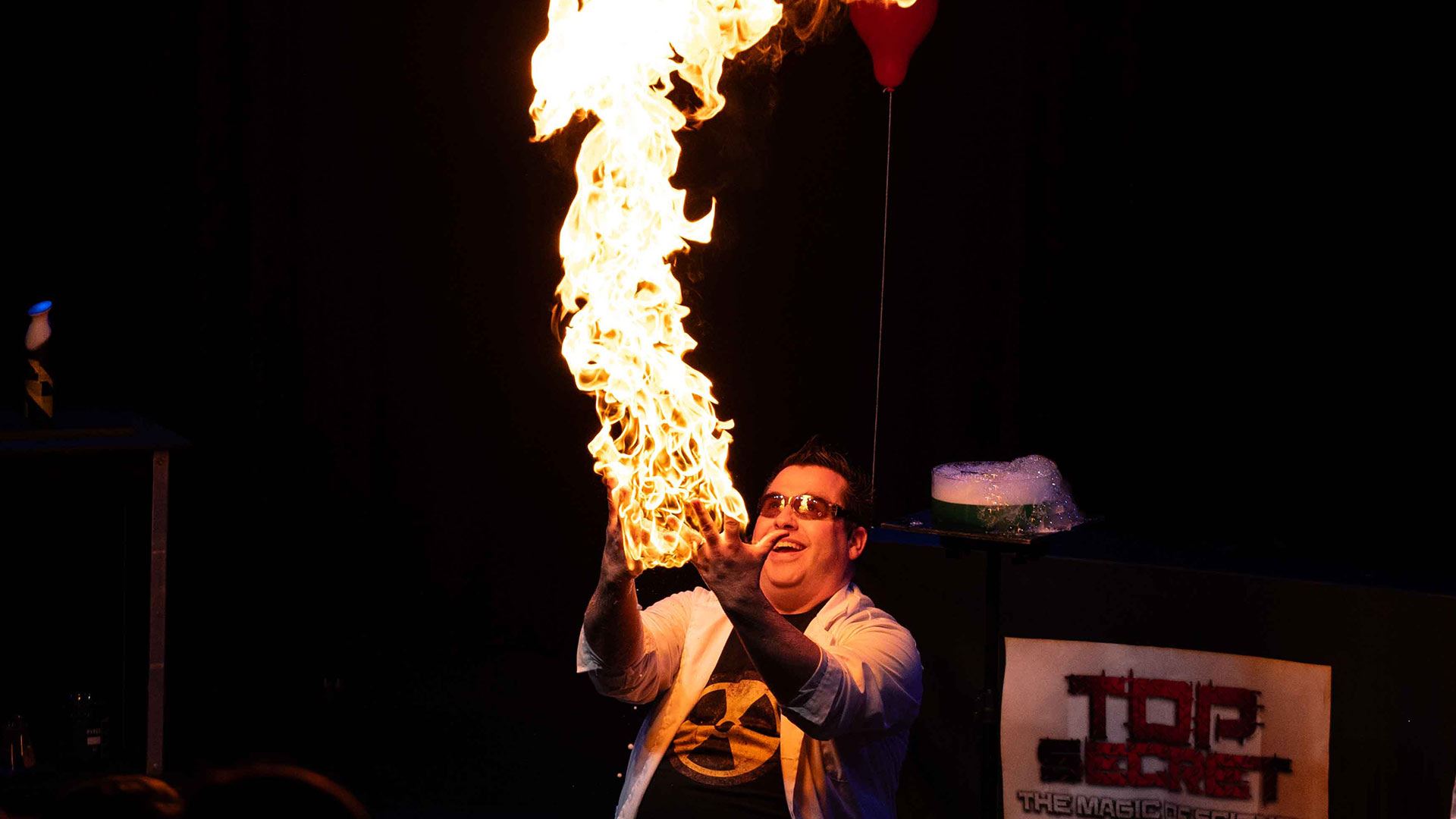 A person in a lab coat creates a tall column of fire during a stage science demonstration.