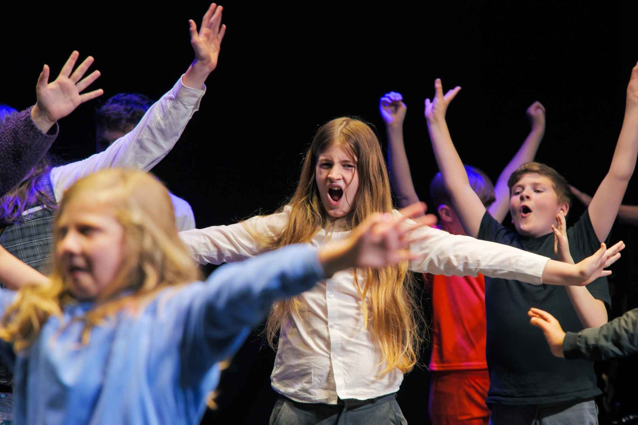 A group of children perform energetically on stage with their arms raised during a theatre workshop or performance. In the centre, a girl with long hair and a white shirt sings or shouts expressively while other children around her strike dramatic poses under colourful stage lighting against a dark background.