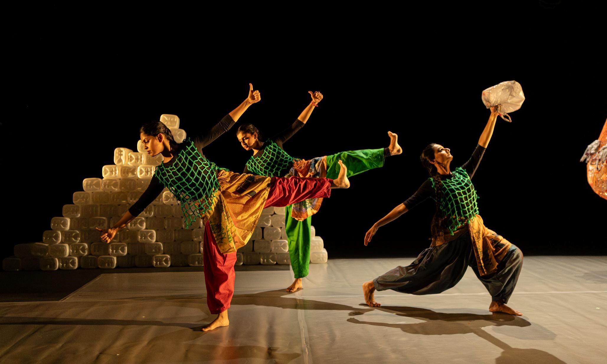 Three dancers perform on a dark stage, balancing in expressive poses while holding plastic containers. They wear colourful costumes with green netting overlays, with stacks of plastic containers forming part of the set behind them.