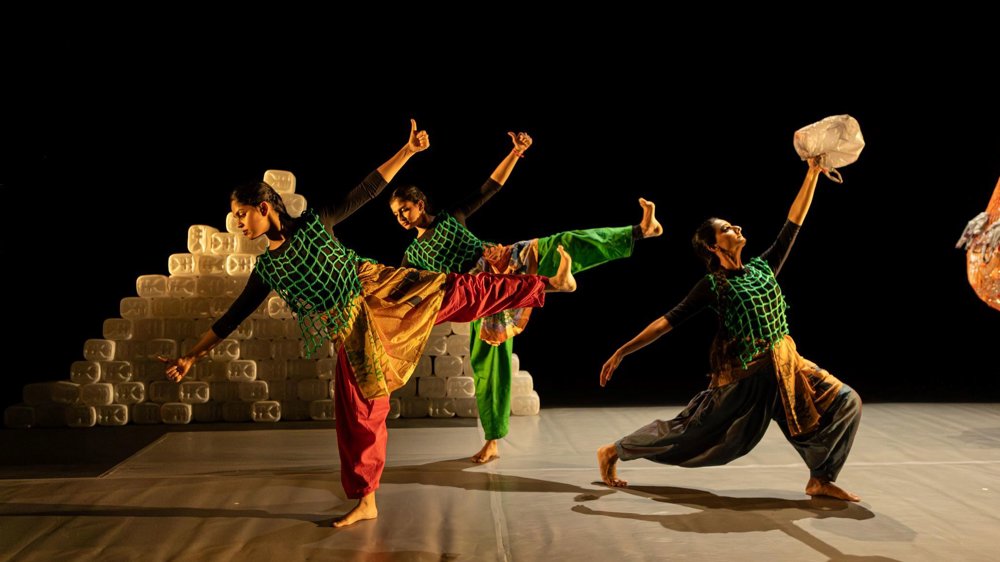 Three dancers perform on a dark stage, balancing in expressive poses while holding plastic containers. They wear colourful costumes with green netting overlays, with stacks of plastic containers forming part of the set behind them.
