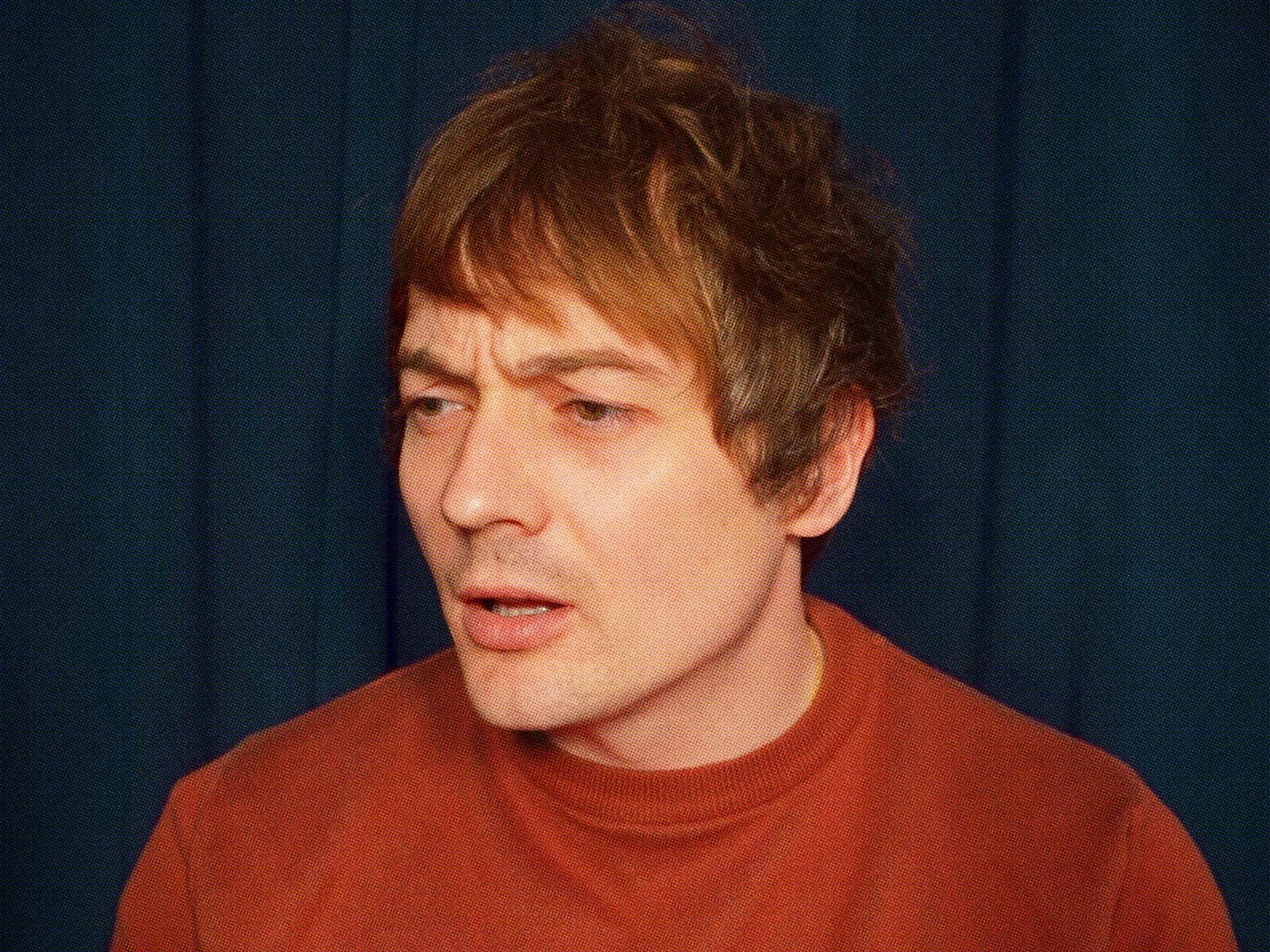 A close-up portrait of a person with short brown hair wearing a red top, looking slightly to the side with a thoughtful expression against a dark blue background.