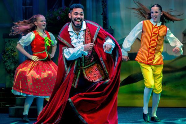 Three pantomime performers dance energetically on stage, led by a smiling man in a red cape, flanked by two performers in colourful storybook costumes, mid-movement under bright stage lighting.