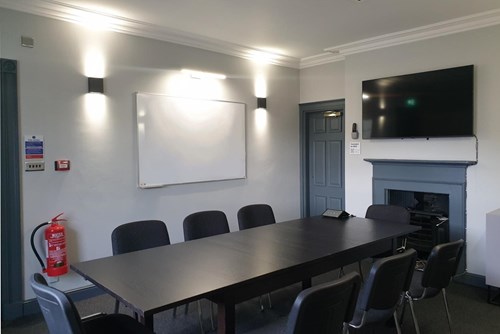 Small Meeting Room at Lawrence Batley Theatre with a large table with 8 chairs around. The walls are a light grey and with a whiteboard on one wall and a TV on another.