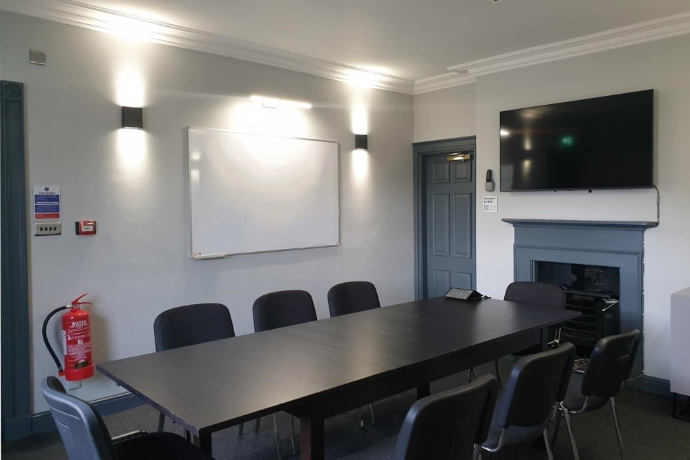 Small Meeting Room at Lawrence Batley Theatre with a large table with 8 chairs around. The walls are a light grey and with a whiteboard on one wall and a TV on another.