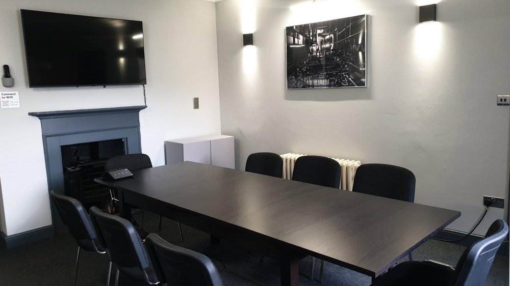 Small Meeting Room at Lawrence Batley Theatre with a large table with 8 chairs around. The walls are a light grey and with a photograph on one wall and a TV on another.