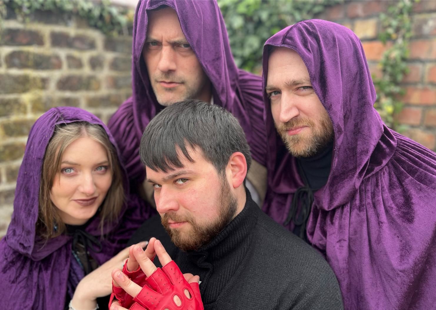 Four people pose closely together outdoors against a brick wall. Three wear deep purple hooded cloaks and look intensely at the camera, while a fourth person in a black jumper and bright red fingerless gloves sits in front with their hands clasped. The group’s dramatic expressions and costumes create a mysterious, fantasy-inspired atmosphere.