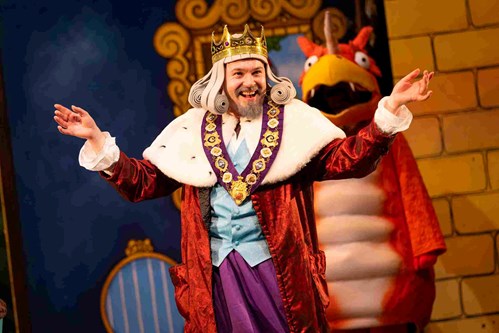 The King is wearing a grey wig with a golden crown on top of his head. His arms are raised showing his red, velvet robe with a big white collar. 