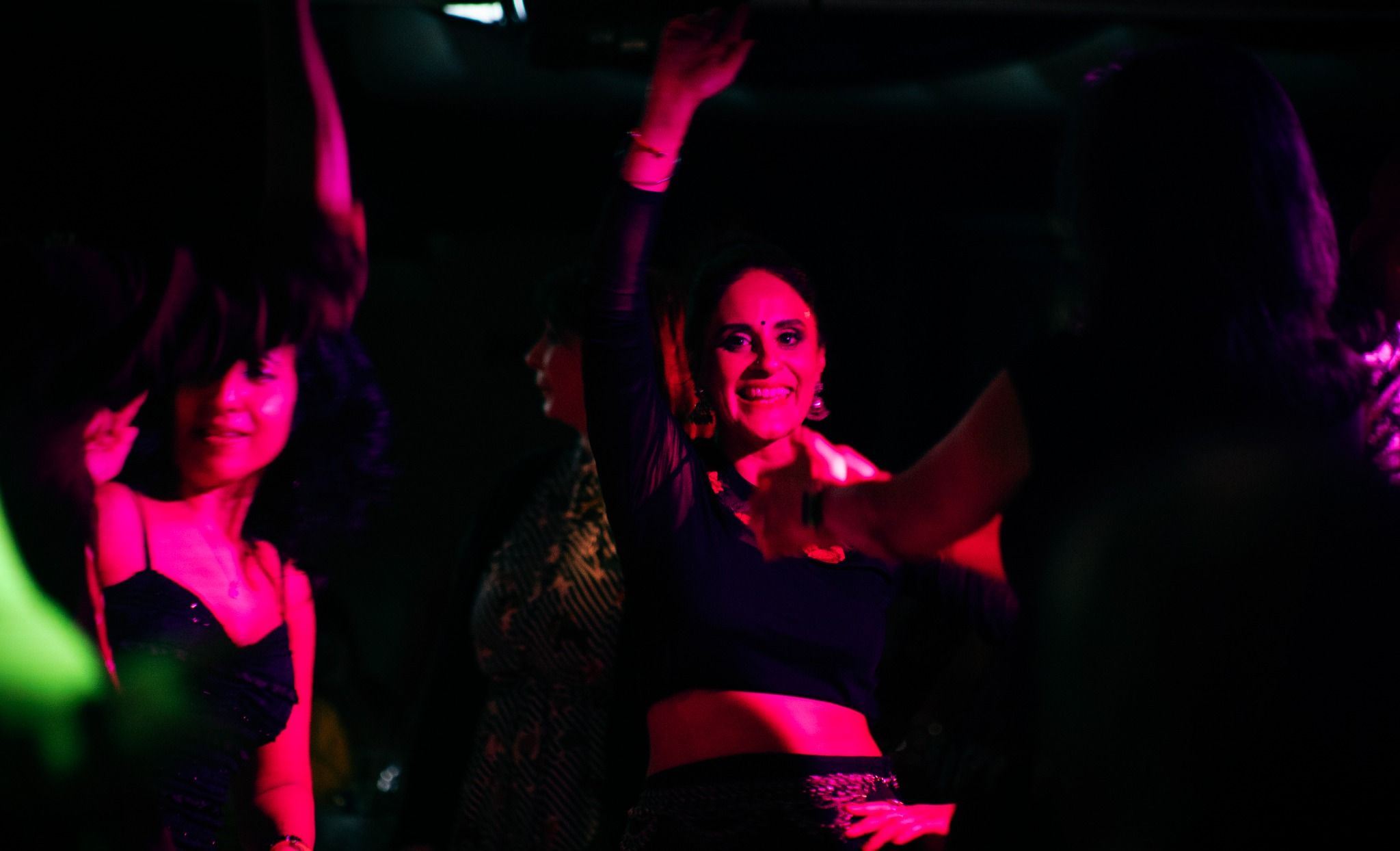 A group of people dance together in a dimly lit venue, illuminated by vibrant pink and purple club lights. One smiling person in the centre raises an arm in the air while others dance around them, creating a lively and energetic party atmosphere.