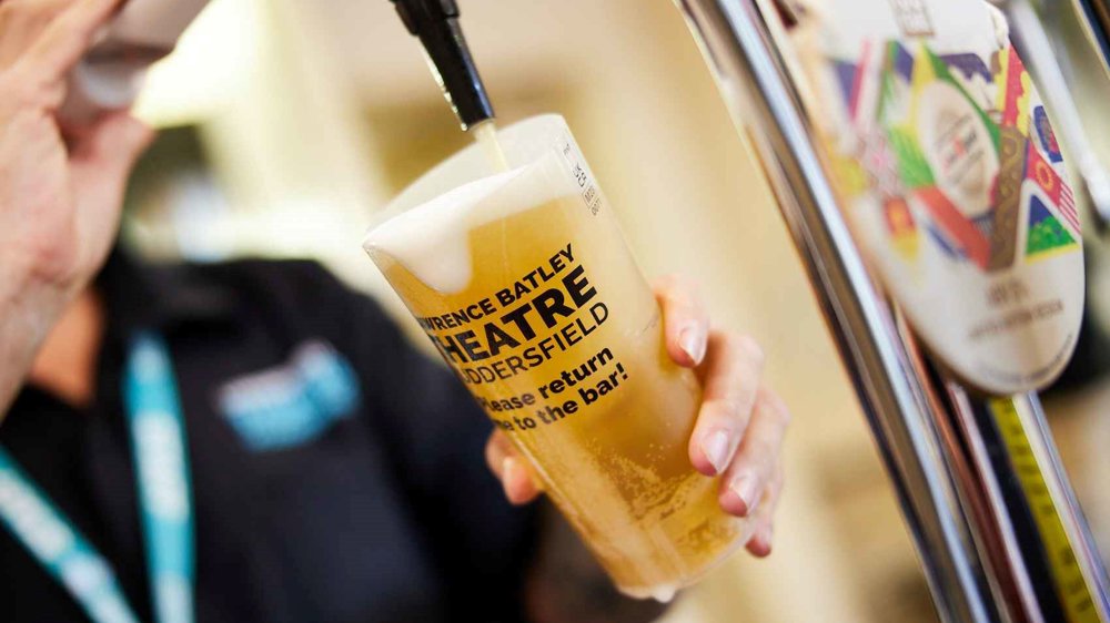 A bartender pours a pint of beer from a tap into a reusable plastic cup branded with "Lawrence Batley Theatre Huddersfield." The bartender wears a black uniform with a lanyard, and the background is blurred, showing a busy bar setting.