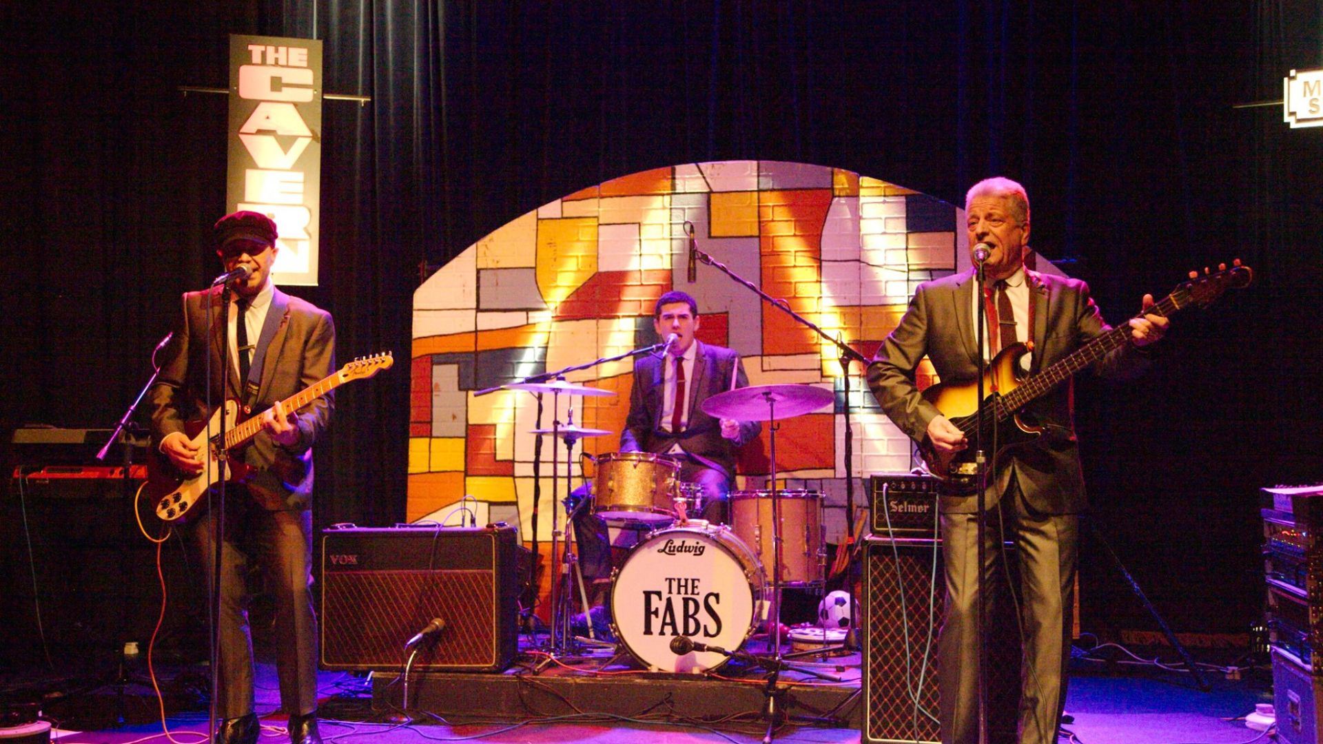 A three-piece band performing on stage in suits, with one playing guitar, one on drums, and one on bass. A drum kit with “The Fabs” logo is centre stage, and behind them is a colourful mural backdrop with the words “The Cavern” lit up to the side.