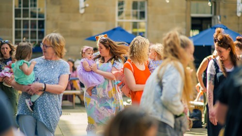 A vibrant outdoor gathering featuring parents and children. A woman in a pastel dress holds a baby in a purple dress, while others interact and socialize in the sunny courtyard setting. Blue umbrellas and a historic building form the backdrop, enhancing the festive atmosphere