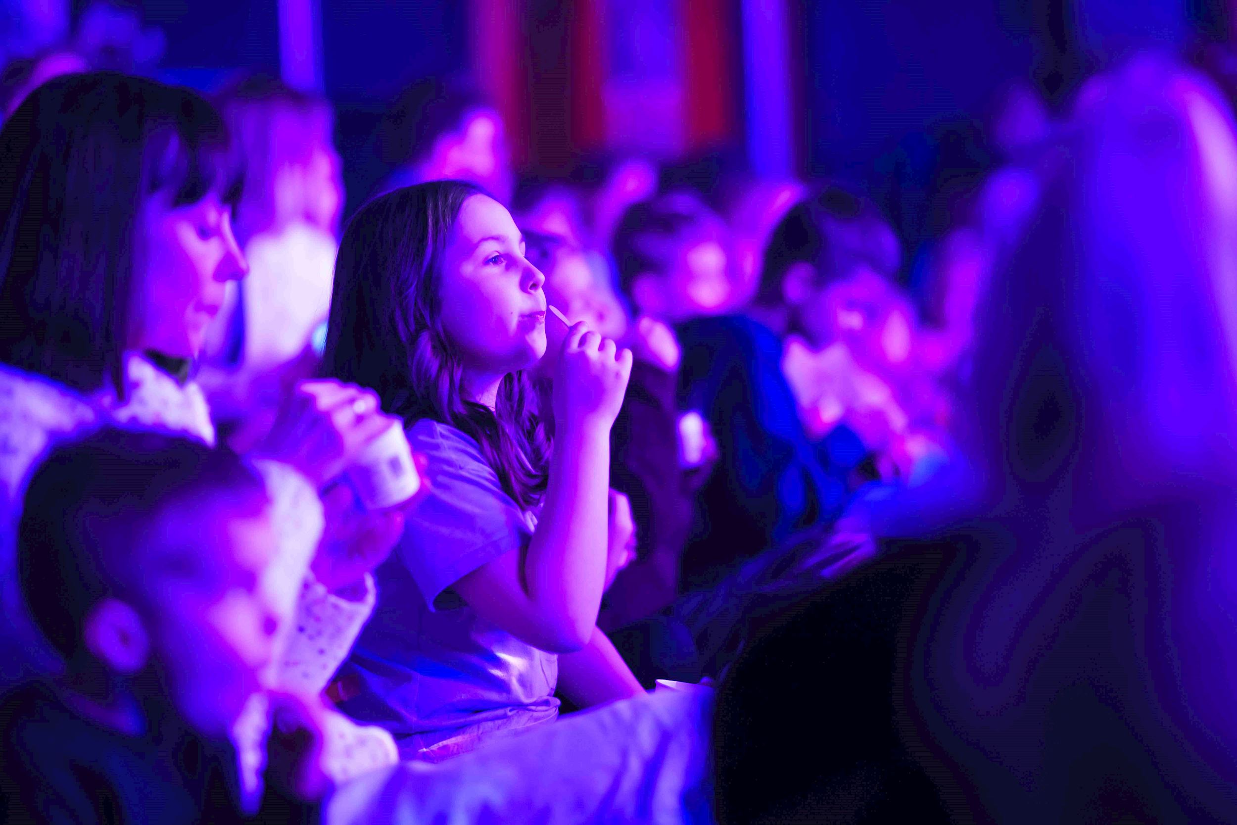 A captivated audience of children and adults sits bathed in vibrant purple lighting, watching a live performance.