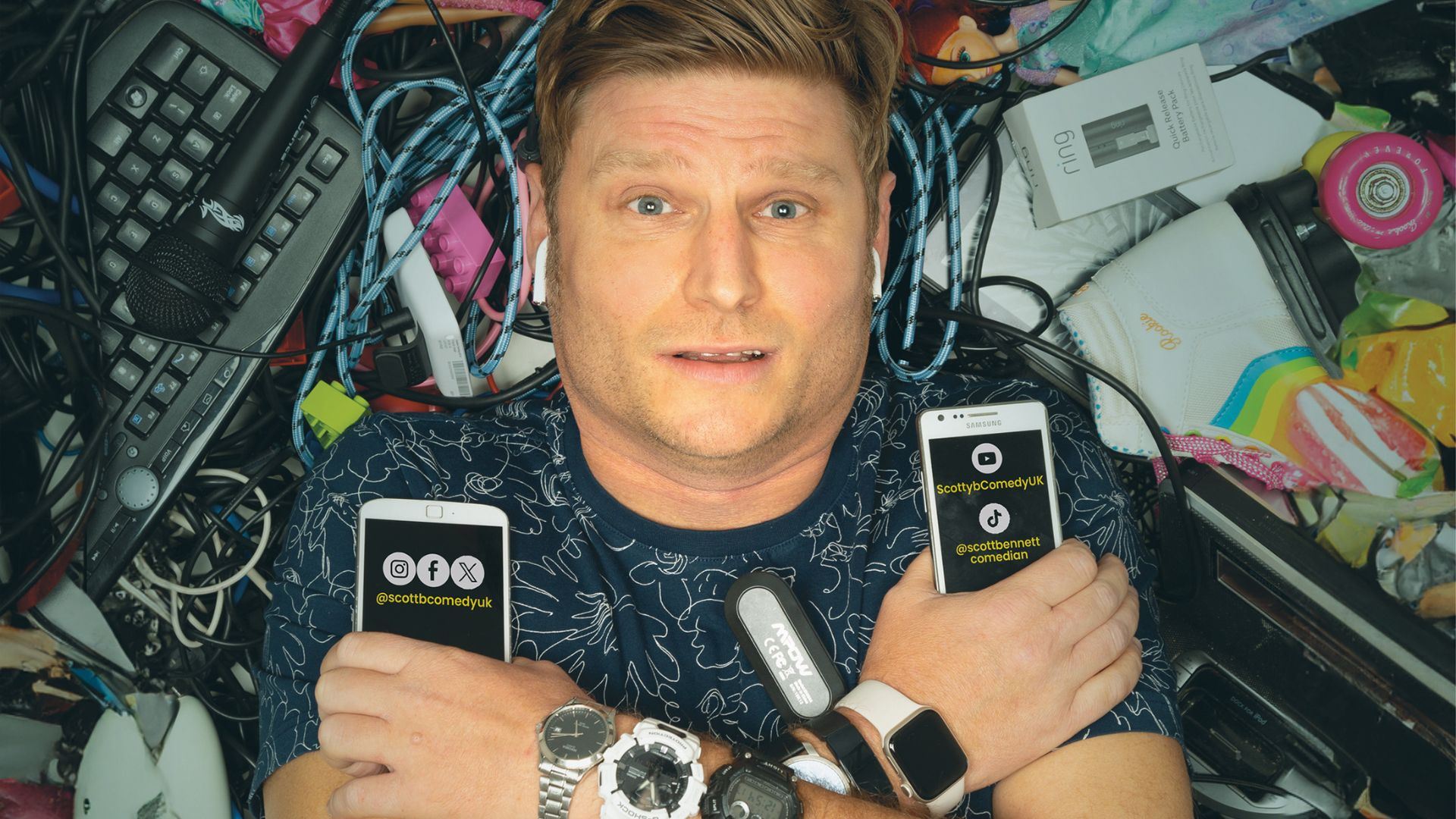 Comedian Scott Bennett lies on his back surrounded by a chaotic tangle of tech gadgets, wires, and miscellaneous items including a microphone, keyboard, smartwatches, and a roller skate. He holds two smartphones displaying his social media handles: @scottbcomedyuk on Instagram, Facebook, and X, and @scottbennettcomedian on TikTok and ScottybComedyUK on YouTube. His expression is humorous and overwhelmed, with earbuds in and multiple smartwatches on his wrists.