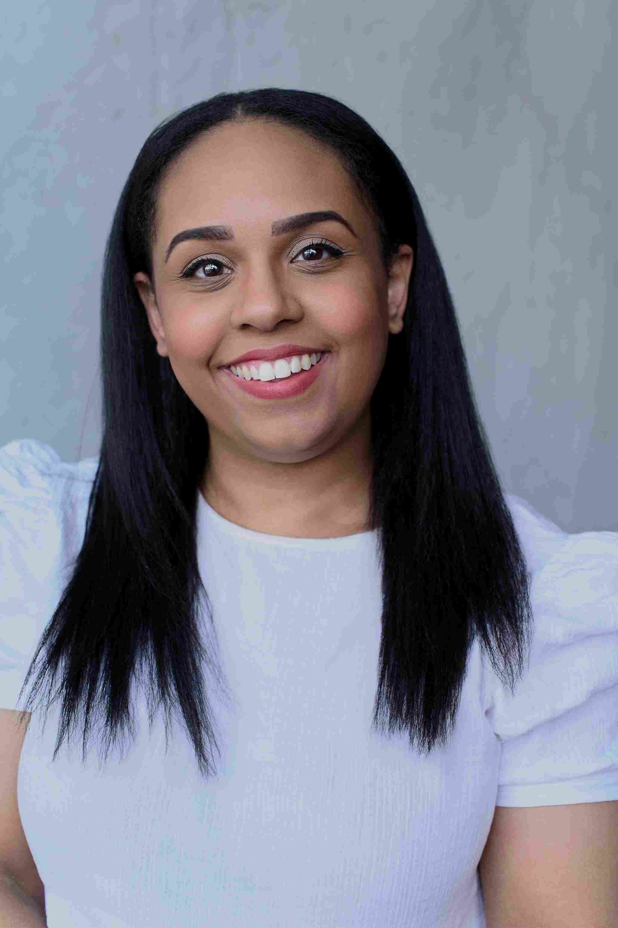 Shorelle Hepkin: Smiling woman with long straight dark hair, wearing a white puff-sleeve top, standing against a pale background.
