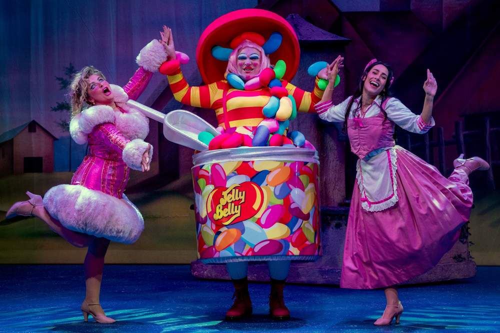 Three pantomime characters pose joyfully on stage, including a performer dressed as a giant Jelly Belly jar, flanked by two performers in pink costumes, all mid-dance against a theatrical backdrop.