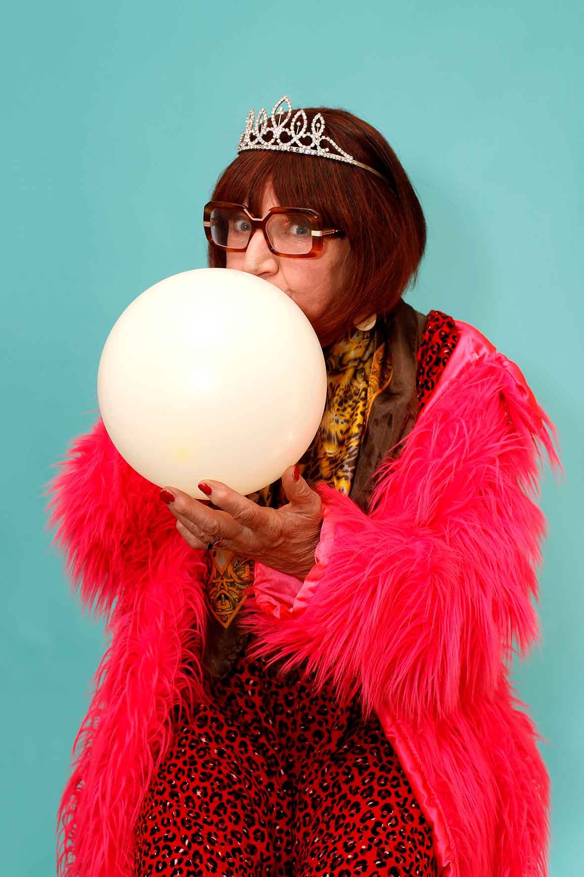 Barbara Nice wearing a sparkly tiara, large glasses, a bright pink faux fur coat, and red leopard-print pants blows up a white balloon against a turquoise background.