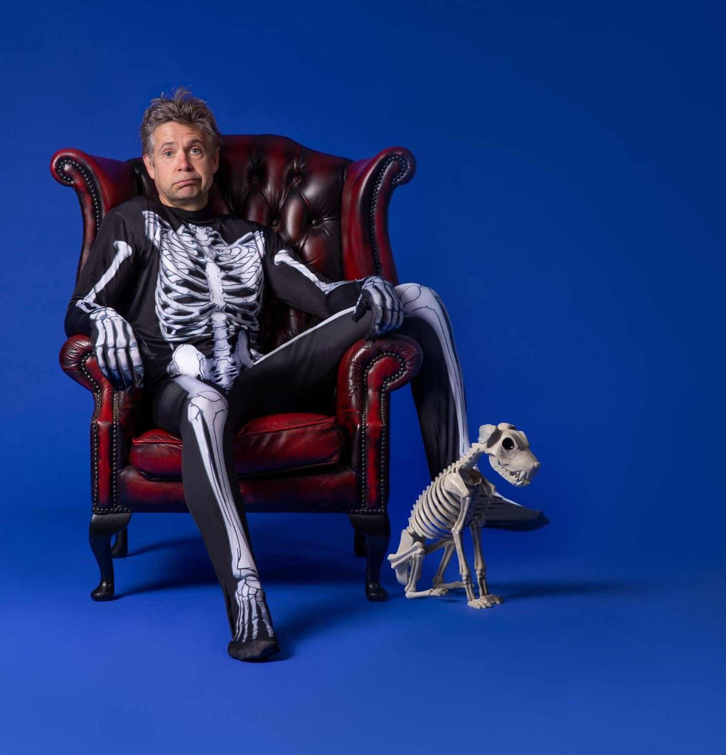 A man wearing a full-body skeleton costume sits in a deep red leather armchair against a solid blue background. He has a slightly surprised expression and tousled hair. Beside him on the floor is a small dog skeleton figure, positioned as if sitting obediently.