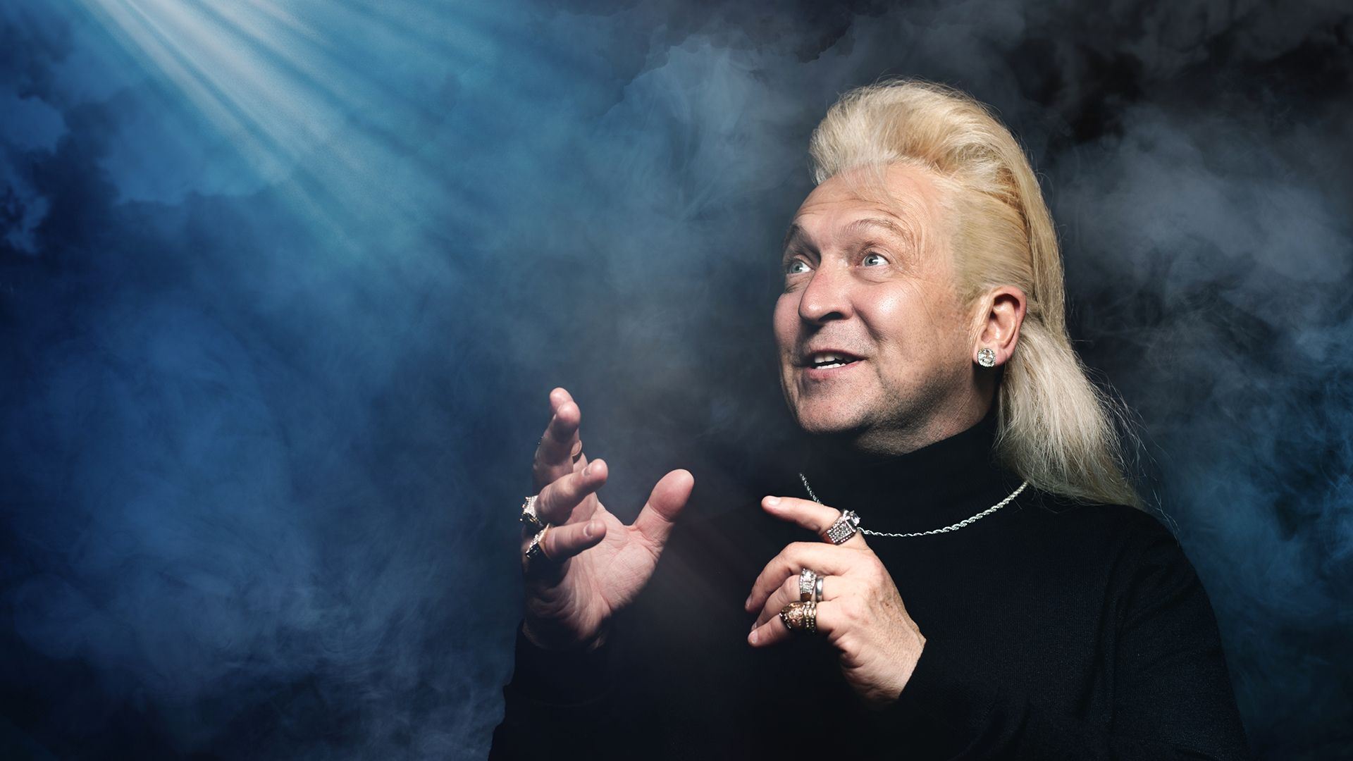 A man with long blond hair and multiple rings gestures expressively while looking upwards. He wears a black turtleneck and silver jewellery, with dramatic lighting and smoky blue background creating a mystical atmosphere