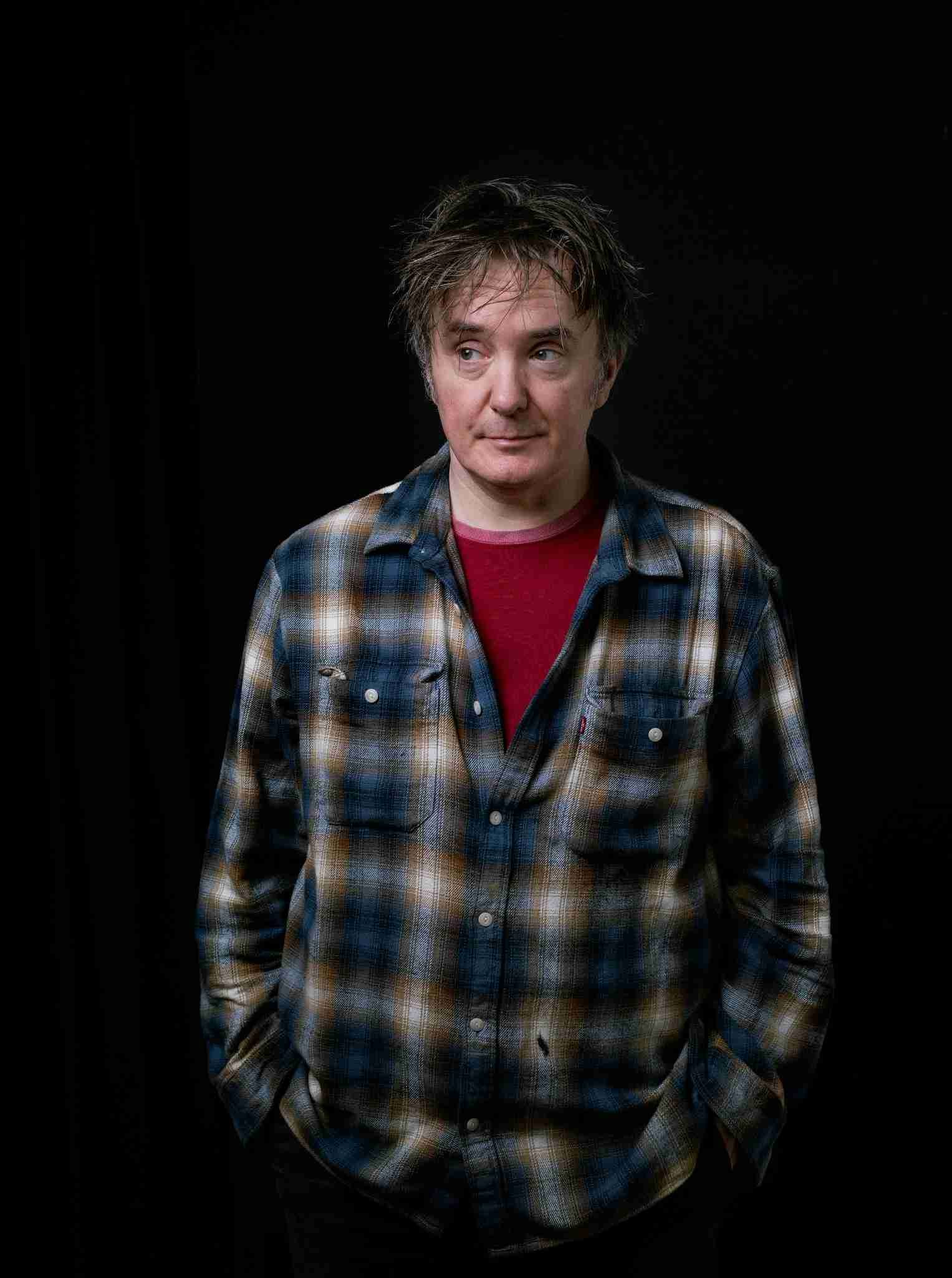 A man stands against a plain black background wearing a blue and brown plaid shirt over a red T-shirt. He faces forward with his hands in his pockets and looks slightly to the side with a thoughtful expression.