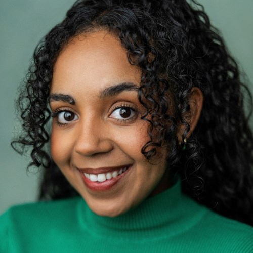 Headshot of Alyce Liburd. She has dark curly hair, wears a green turtleneck, and smiles warmly at the camera.